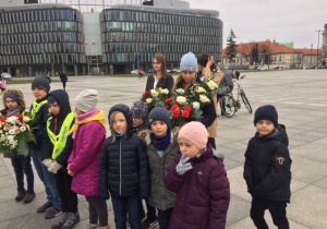 Uczniowie naszej szkoły, podczas uroczystości, stoją na placu z kwiatami.