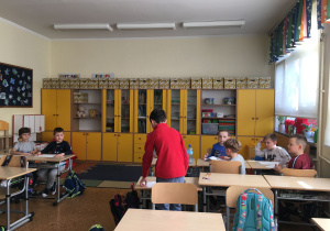 Adam, stojąc przy ławkach uczniów, tłumaczy i pokazuje, jak składać papier.