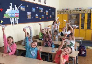 Uczniowie siedzą w ławkach i pokazują swoje papierowe łódki.