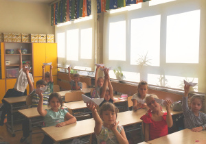 Uczniowie siedzą w ławkach i pokazują swoje papierowe łódki.
