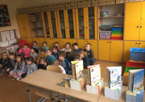 Uczniowie siedzą na dywanie i biorą udział w zagadkach, które są częścią spotkania.