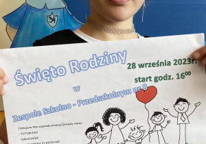 Uśmiechnięta trzecioklasistka prezentuje plakat informujący o szkolno-przedszkolnym wydarzeniu z okazji Dnia Rodziny.