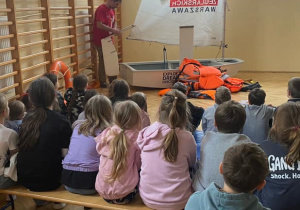 Uczniowie słuchają osoby prowadzącej, która opowiada o zachowaniu bezpieczeństwa nad wodą.