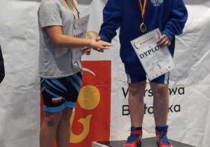 Karol, podczas dekoracji medalami, stoi na podium i przyjmuje gratulacje od uczestnika zapasów.