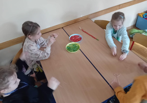 Uczniwoie siedzą przy ławkach i nawlekają na sznurki kolorowe dziesiątki z makaronu.