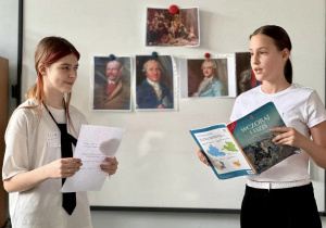 Dwie uczennice stoją przy tablicy, na której wisza zdjęcia Króla Stanisława Augusta Poniatowskiego i prezentują swój pokaz.