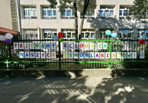 Napis "Trzymamy kciuki za naszych ósmoklasistów" ułożony z kolorowych liter i ozdobiony balonami wisi na ogrodzeniu szkoły.