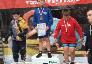 Karol stoi na podium podczas dekoracji medalami. W rękach trzyma dyplom.