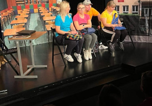 Spektakl trwa. Aktorzy siedzą na krzesłach. Scenografia pokazuje wnętrze sali klasowej i wejście do szkoły.