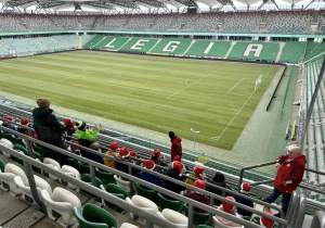 Uczniowie siedzą na trybunie stadionu i słuchają historii klubu. W tle widać napis "Legia" ułożony z krzesełek w kolorze białym.