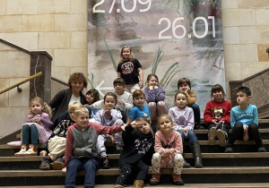 Grupowe zdjęcie pierwszoklasistów. Dzieci razem z Panią Iwoną siedzą na kamiennych schodach. W tle widać plakat z informacją o wystawie Józefa Chełmońskiego i czasie jej trwania.