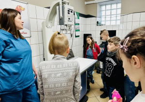 Dzieci stoją w gabinecie. Pani doktor wyjaśnia do czego służą fartuchy wypełnione ołowiem.