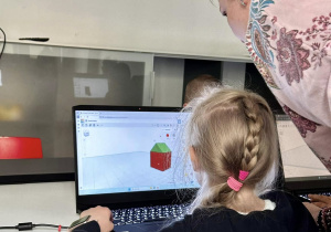 Uczennica siedzi przy komputerze i słucha wskazówek, jak poprawnie wykonać, a potem zapisać do druku swój model 3D.