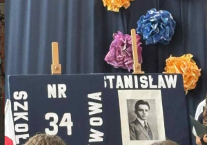 Na tle kotary, do której przypięta jest dekoracja widać atrybuty szkoły, tarczę oraz zdjęcie patrona szkoły - Stanisława Dubois.