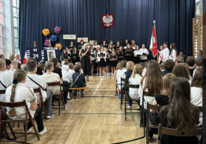 Pani Dyrektor przemawia do zebranych uczniów i rodziców, w tle stoi szkolny chór, który z okazji rozpoczęcia roku szkolnego przygotował program artystyczny.
