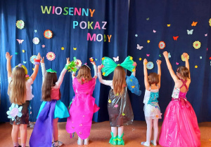 Dziewczynki prezentują piękne i kolorowe stroje stojąc na tle napisu dekoracyjnego "Wiosenny Pokaz Mody".