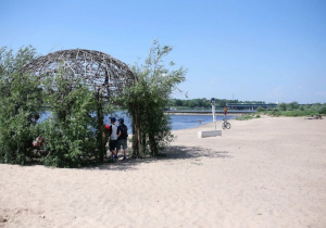 Uczniowie stoją na plaży nad Wisłą pod zadaszeniem wykonanym z wikliny.