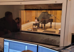 Reżysernia dźwiękowa - dzieci w roli inżynierów dźwięku za mikserem audio i przed komputerem do rejestracji nagrania jednej z artystek klasowych - Julii. Za ścianą widać studio nagrań z fortepianem w trakcie nagrania.