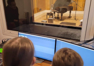 Reżysernia dźwiękowa - dzieci w roli inżynierów dźwięku za mikserem audio i przed komputerem do rejestracji nagrania jednej z artystek klasowych - Zofii. Za ścianą widać studio nagrań z fortepianem w trakcie nagrania.