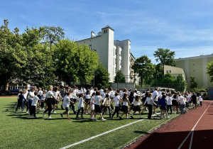 Uczniowie i nauczyciele, ubrani w białe bluzki, tańczą Poloneza na szkolnym boisku.