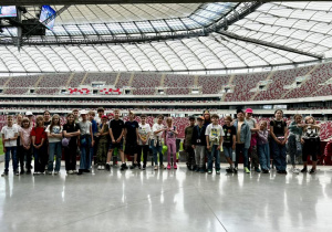 Uczniowie stoją przy krzesełkach kibiców pozując do zdjęcia grupowego. W tle widać dach stadionu.