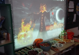 Pod tablicą na której wyświetlana jest "Piosenka na Halloween" stoi stół z przekąskami.