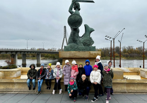 Drugoklasiści stoją pod pomnikiem Syreny i pozują do grupowego zdjęcia.