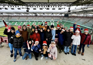 Uczniowie siedzą na trybunie stadionu i słuchają historii klubu. W tle widać napis "Legia" ułożony z krzesełek w kolorze białym.