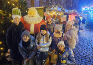 Uczniowie znajdują się na rynku miasta i pozują z Mikołajem do grupowego zdjęcia.