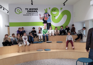 Dzieci siedzą na dużych stopniach i słuchają Pana prowadzącego zajęcia.