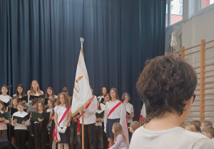 Szkolny chór oraz starsi uczniowie trzymający sztandar szkoły.