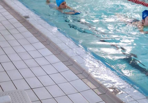 Uczniowie pływają w basenie. Dzieci używają desek do pływania.