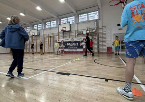 Uczniowie stoja w sali gimnastycznej i słuchają zasad gry zespołowej "Palant".