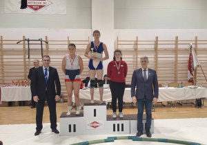 Nasza uczennica, Katarzyna Sidorowicz, stoi na podium wsród pozostałych zawodniczek, po dekoracji medalami i w ręku trzyma puchar.