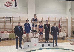 Nasza uczennica, Katarzyna Sidorowicz, stoi na podium wsród pozostałych zawodniczek, po dekoracji medalami i w ręku trzyma puchar.