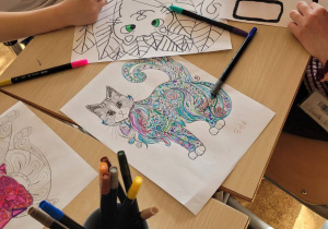 Uczniowie siedzą w ławkach i kolorują kolorowanki z wizerunkiem kota.