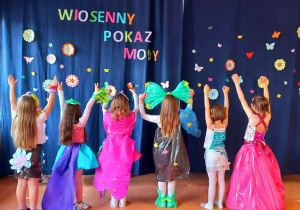 Dziewczynki prezentują piękne i kolorowe stroje stojąc na tle napisu dekoracyjnego "Wiosenny Pokaz Mody".