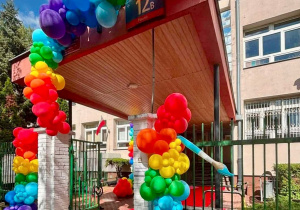 Kolorowe balony ozdabiają wejscie do szkoły, na uczniów i i ch rodziców czeka czerwony dywan, który leży w wejściu do szkoły.