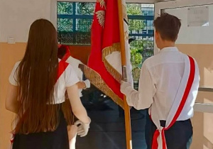 Starsi uczniowie, w strojach galowych, wchodzą do sali gimnastycznej. W rękach trzymają sztandar szkoły.
