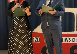 Na zdjęciu widać dwoje nauczycieli stojących na scenie podczas występu z okazji 120-lecia szkoły. Po lewej stoi kobieta, ubrana w długą czarną sukienkę w białe kropki oraz ciemną marynarkę. Trzyma mikrofon i zieloną teczkę z tekstem wystąpienia. Po prawej stoi mężczyzna, w ciemnym garniturze, białej koszuli i krawacie. Również trzyma zieloną teczkę. W tle znajduje się duża dekoracja przypominająca autobus, z widocznym fragmentem napisu oraz numerem "120".