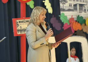 Na zdjęciu widać kobietę stojącą na scenie. Kobieta trzyma mikrofon i przemawia. Za nią znajduje się duży czerwony znak z literą "A" oraz tabliczka z napisem "Warszawa". W tle widoczny jest fragment starej architektury oraz kolorowe, wycinane elementy w kształcie liści.