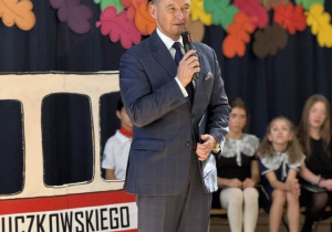Na zdjęciu widać mężczyznę stojącego na scenie. Pan Burmistrz trzyma mikrofon i przemawia. Za nim znajduje się fragment dekoracji w kształcie autobusu. W tle widoczny jest fragment starej architektury oraz kolorowe, wycinane elementy w kształcie liści.