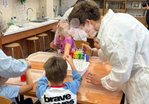 Na zdjęciu widać dzieci uczestniczące w zajęciach laboratoryjnych, siedzące przy dużych stołach w jasnej, przestronnej sali. Obok nich stoją starsi uczniowie, którzy pomagali dzieciom w wykonaniu zadania.